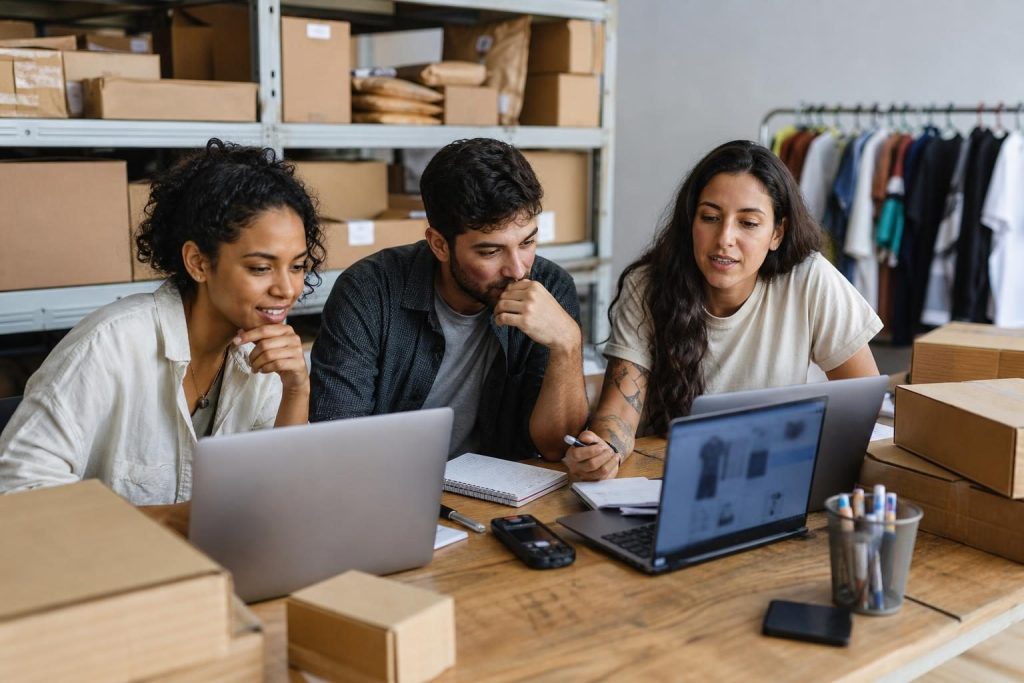 Equipe brasileira de e-commerce revisando páginas de produto criadas com apoio de IA em um escritório com estoque ao fundo